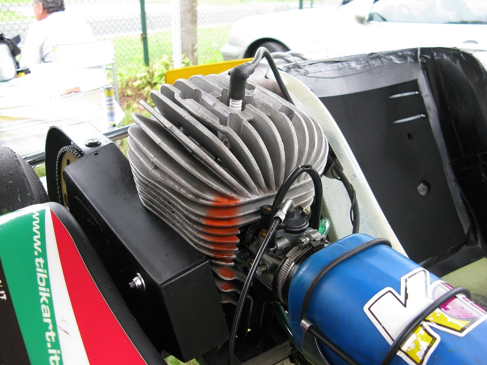 Close view of an air-cooled kart engine with a finned aluminium cylinder head, a spark plug with its cap on top and a carburettor with a blue intake silencer on the right