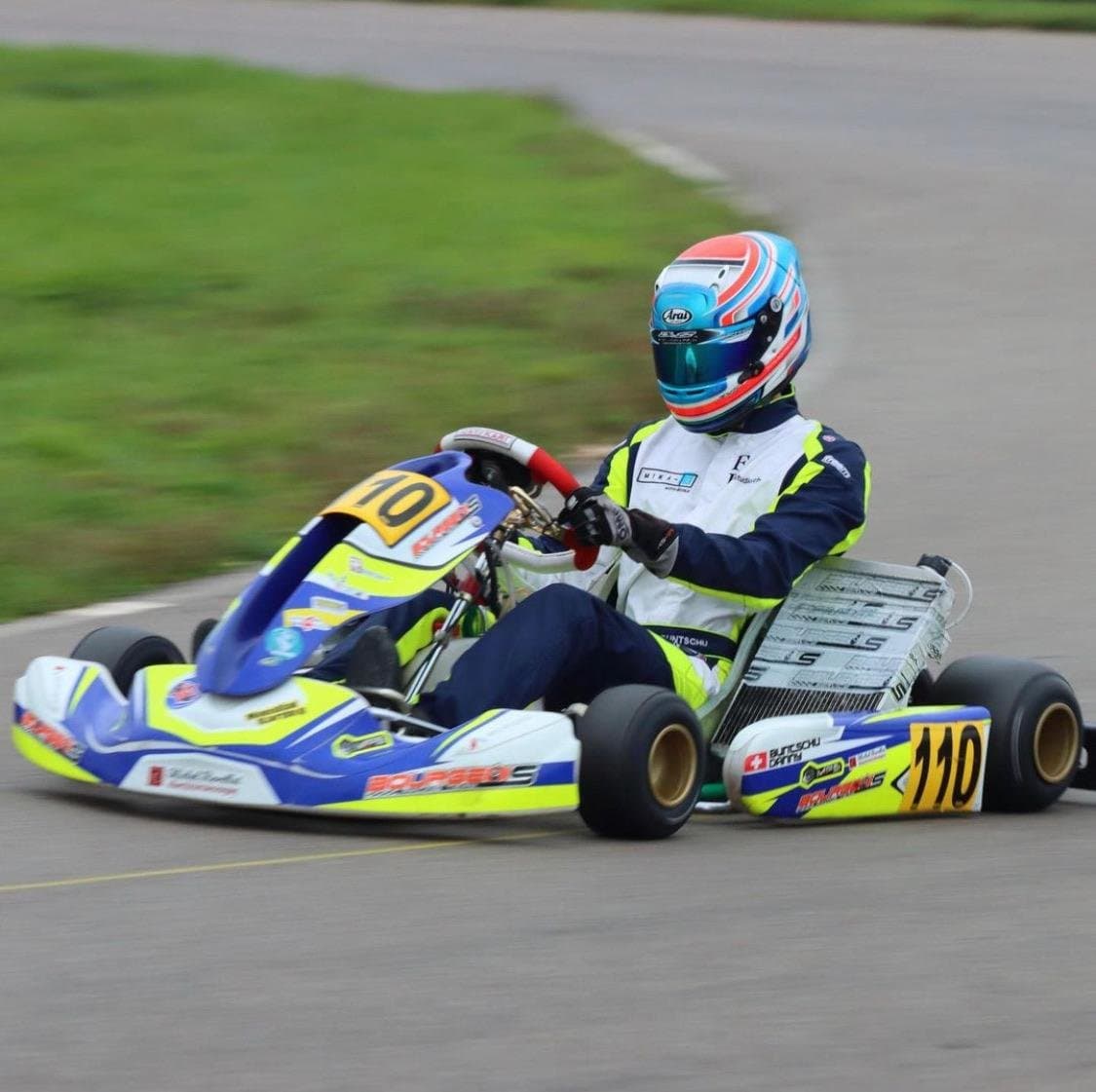 A KZ2 kart, number 110, cornering at speed on grey tarmac with the grass and track edge blurred by the panning shot