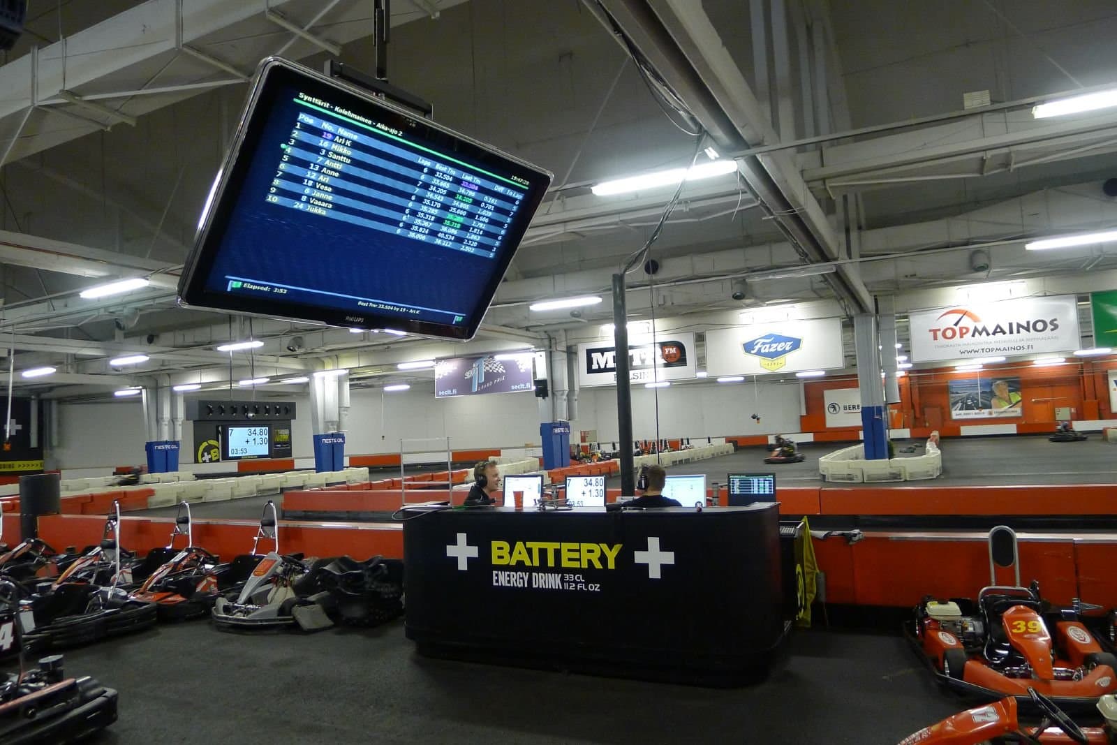 Ceiling mounted monitor showing a race results table with positions, laps, best times and last times above a race control desk at an indoor kart track
