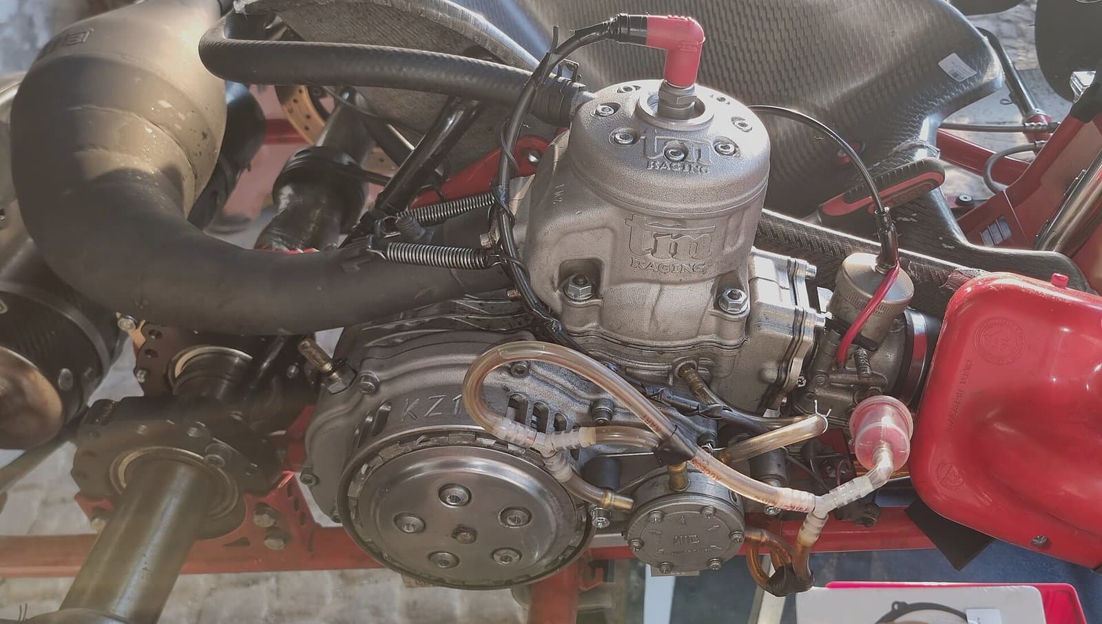 Side view of a TM KZ10 two-stroke kart engine on a kart chassis, with the big black exhaust header and expansion chamber curving away on the left and a red fuel tank on the right