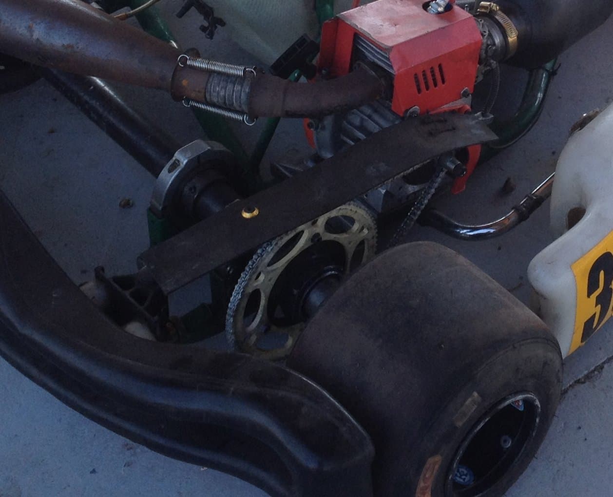 Rear left of a racing kart showing the large gold rear sprocket on the axle and the chain running up to the engine behind a black chain guard
