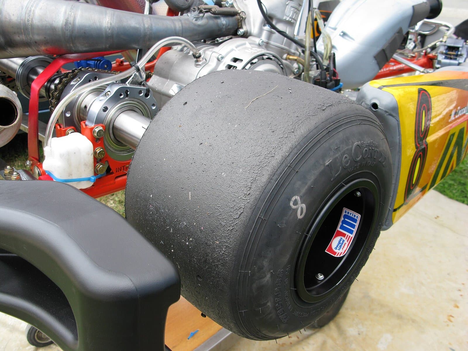 Close view of a worn LeCont slick tire on a kart rear wheel, with the axle, chain and engine behind it