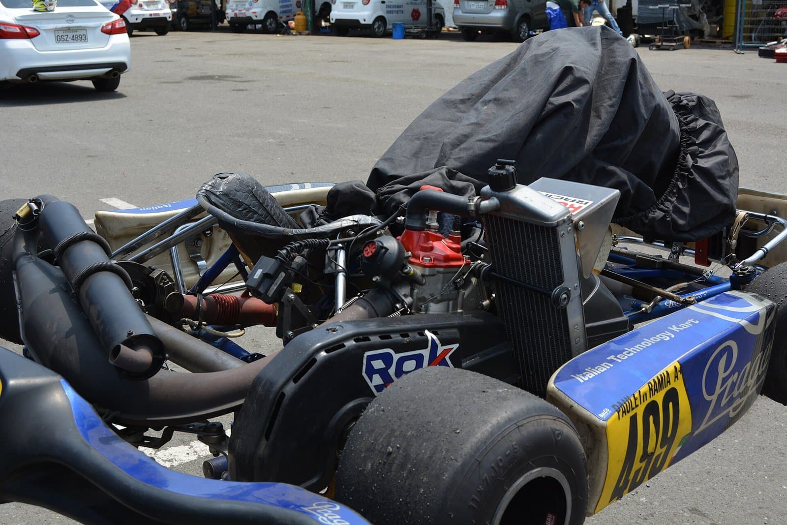 Side view of a racing kart with a black aluminium radiator beside the seat, a silver top tank with a cap, a coolant hose running to the red cylinder head and the exhaust pipe on the left