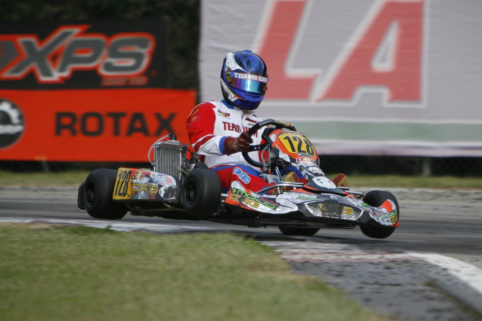 A kart turning into a corner on a paved circuit, the driver braced behind the wheel while a wheel starts to lift off the asphalt.