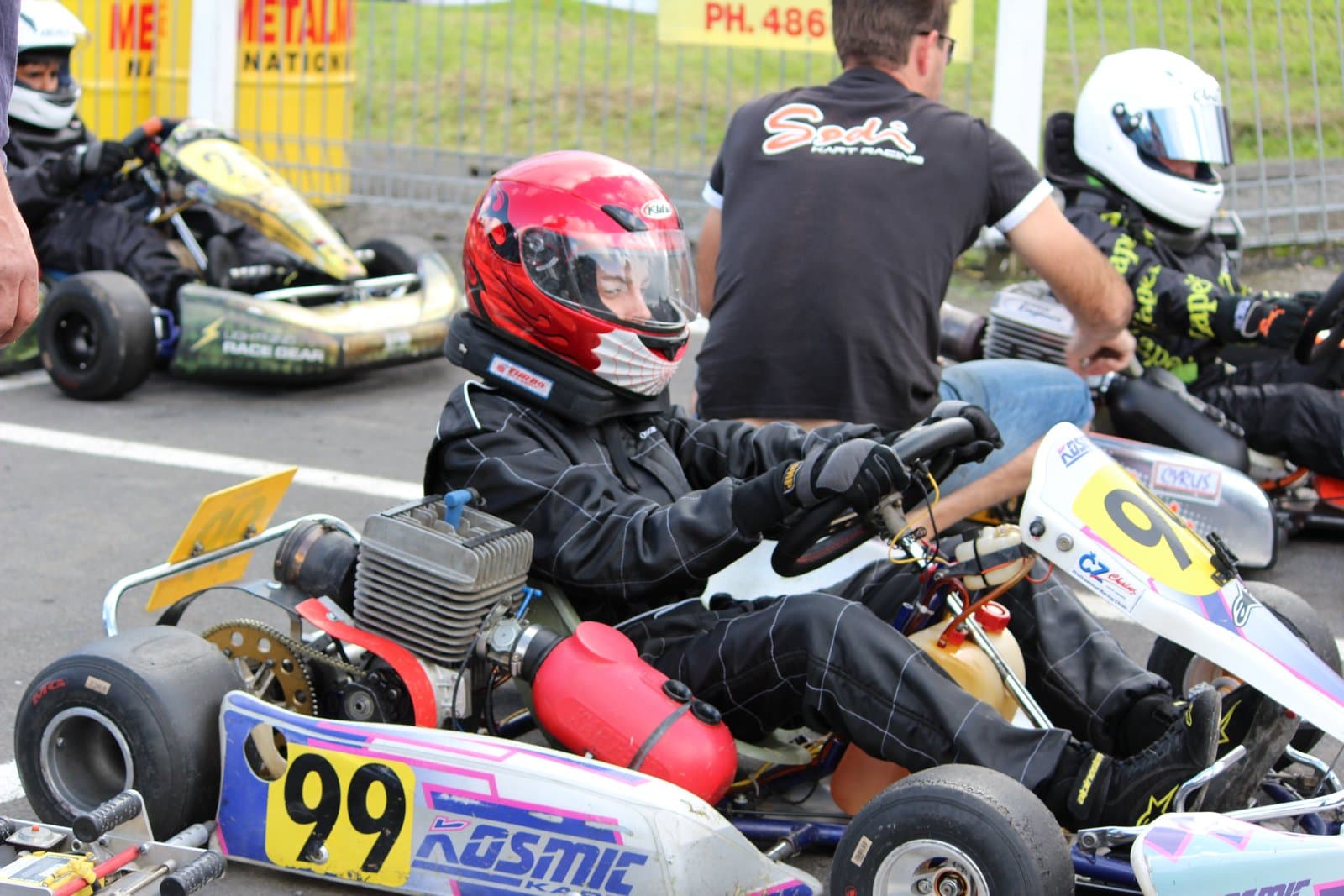 A young driver in a red helmet and black race suit sits in a kart on the grid, hands on the wheel, waiting for the start with other karts around
