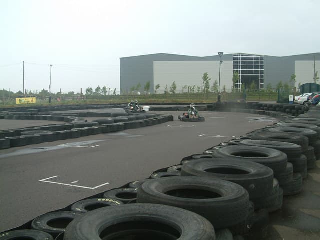 Karts on a damp kart circuit in the rain, with tyre walls on the right and a grey sky